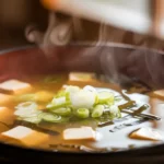 A close-up of a steaming bowl of Japanese soup with tofu, green onions, and seaweed in a rich miso broth.